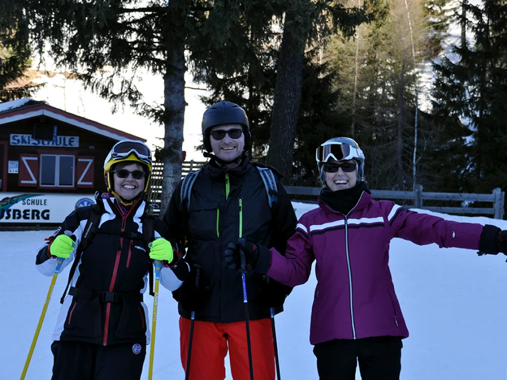 Drei Personen posieren mit ausgebreiteten Armen vor einer Hütte.